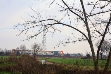 Electricity pylon and an abandoned building in the distance framed by bare trees in a field on a cloudy day in winter