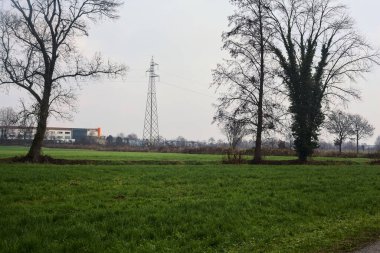 Electricity pylon and an abandoned building in the distance framed by bare trees in a field on a cloudy day in winter