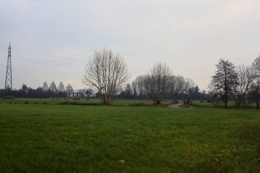 Asphalt trail bordered by fields and a stream of water with a bridge crossing it next to a group of bare trees on a cloudy day