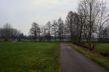 Asphalt trail bordered by fields and a stream of water with a bridge crossing it next to a group of bare trees on a cloudy day