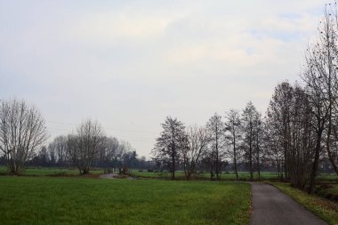 Asphalt trail bordered by fields and a stream of water with a bridge crossing it next to a group of bare trees on a cloudy day