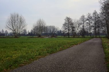 Asphalt trail bordered by fields and a stream of water with a bridge crossing it next to a group of bare trees on a cloudy day
