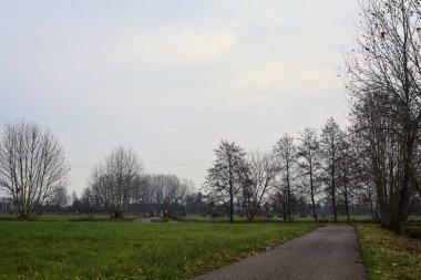 Asphalt trail bordered by fields and a stream of water with a bridge crossing it next to a group of bare trees on a cloudy day
