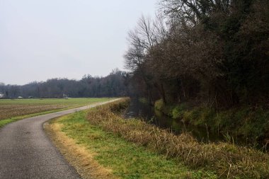 Asphalt trail bordered by a cultivated field and a forest next to a stream of water on a cloudy day in the italian countryside