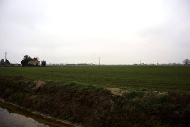 Abandoned country house in a field next to a road and a stream of water on a cloudy day in the italian countryside