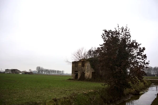 Abandoned country house covered by plants in a cultivated field next to a road and a stream of water on a cloudy day in the italian countryside
