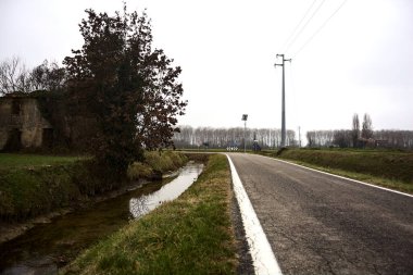 Abandoned country house covered by plants in a cultivated field next to a road and a stream of water on a cloudy day in the italian countryside