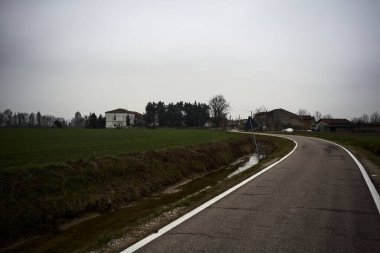Country road bordered by fields with a group of houses in the distance on a cloudy day in the italian countryside
