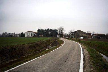 Country road bordered by fields with a group of houses in the distance on a cloudy day in the italian countryside