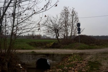 Crossing between a dirt trail and a paved one next to a stream of water on a cloudy day in the italian countryside