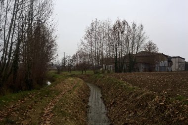 Trail bordered by irrigation channel and bare trees on a cloudy day in the italian countryside in winter