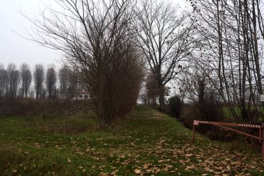 Dirt path bordered by bare plants and trees blocked by a bar next to a field on a cloudy day in the italian countryside