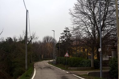 Country road bordered by wooden pylons and fields with a group of houses on a cloudy day
