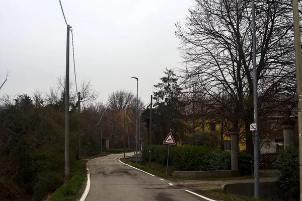 Country road bordered by wooden pylons and fields with a group of houses on a cloudy day