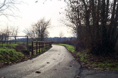Paved trail bordered by a railing and a stream of water between fields and bare trees in the italian countryside