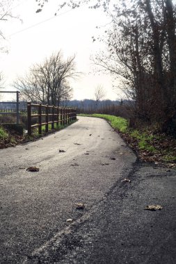 Paved trail bordered by a railing and a stream of water between fields and bare trees in the italian countryside