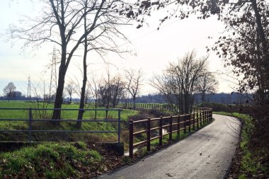 Paved trail bordered by a railing and a stream of water between fields and bare trees in the italian countryside
