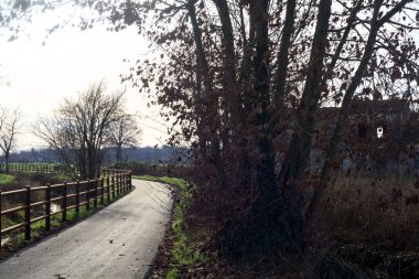Paved trail bordered by a railing and a stream of water between fields and bare trees in the italian countryside