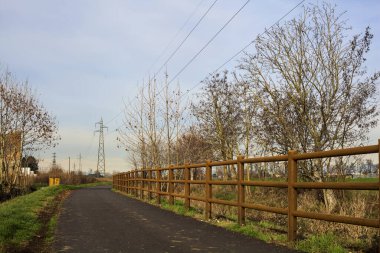 Paved trail bordered by a railing and a stream of water between fields and bare trees in the italian countryside