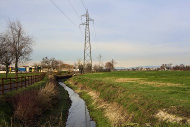 Paved trail bordered by a railing and a stream of water between fields and bare trees in the italian countryside