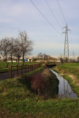Paved trail bordered by a railing and a stream of water between fields and bare trees in the italian countryside