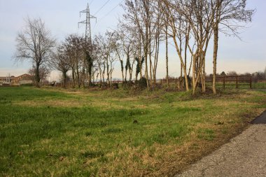 Paved trail bordered by a railing and a stream of water between fields and bare trees in the italian countryside