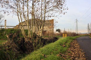 Paved trail bordered by a railing and a stream of water between fields and bare trees in the italian countryside