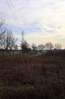Paved trail bordered by a railing and a stream of water between fields and bare trees in the italian countryside