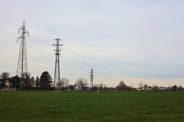 Country road between fields with bare trees and electricity pylons with over head cables on a cloudy day