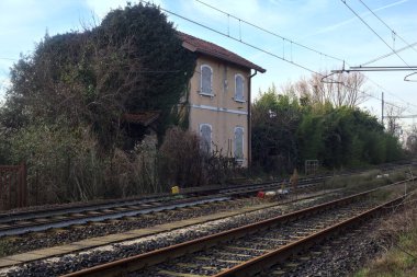 Abandoned building covered by ivy by the edge of a railroad track on a cloudy day in the italian countryside