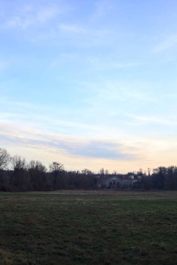 Çıplak ağaçlarla dolu patikanın yanındaki çayır ve gün batımında parkta bir köşk.