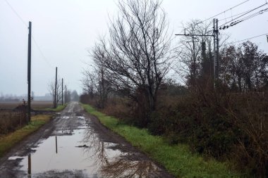 Dirt road with puddles bordered by a field and trees by the edge of a railroad track on an embankment on a cloudy day in the italian countryside