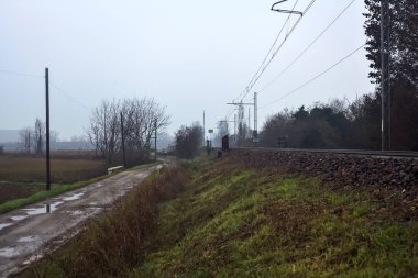 Dirt road and a bridge next to an embankment and a railroad track on a cloudy day in the italian countryside in winter
