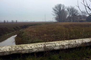 Stream of water between cultivated fields seen from a bridge on a cloudy day in the italian countryside in winter