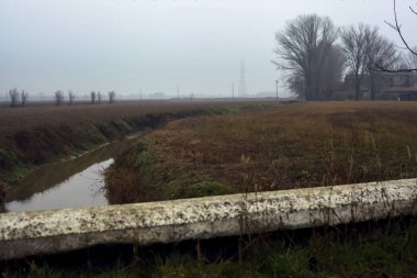 Stream of water between cultivated fields seen from a bridge on a cloudy day in the italian countryside in winter