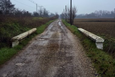 Dirt road and a bridge next to an embankment and a railroad track on a cloudy day in the italian countryside in winter