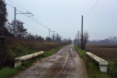 Dirt road and a bridge next to an embankment and a railroad track on a cloudy day in the italian countryside in winter