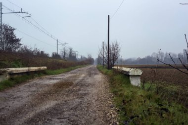 Dirt road and a bridge next to an embankment and a railroad track on a cloudy day in the italian countryside in winter