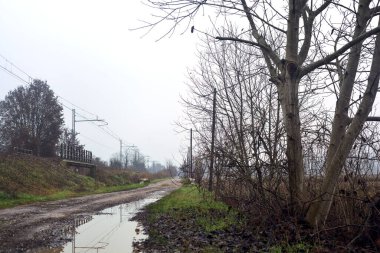 Dirt road and a bridge next to an embankment and a railroad track on a cloudy day in the italian countryside in winter