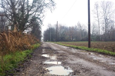 Dirt road with puddles bordered by a field and trees by the edge of a railroad track on an embankment on a cloudy day in the italian countryside