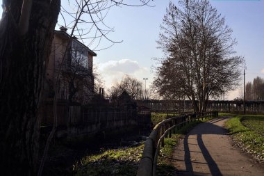 Paved trail bordered by bare trees next to a stream of water and a group of houses on a sunny day in the italian countryside