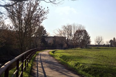 Paved trail bordered by bare trees next to a stream of water and a group of houses on a sunny day in the italian countryside