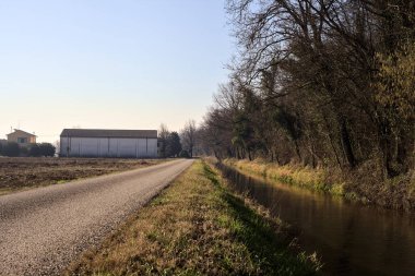 Road and a stream of water next to a forest and a field in the italian countryside in winter