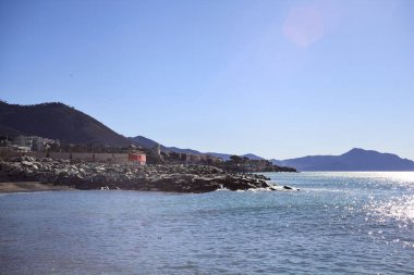 Sahilde dalgalı sular ve güneşli bir günde arka planda uzanan bir şehir.