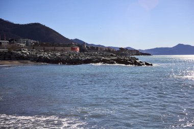 Sahilde dalgalı sular ve güneşli bir günde arka planda uzanan bir şehir.