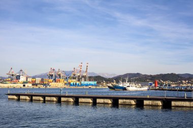 Gün batımında bir körfezde, uzak mesafeden görülen bir nakliye deposu ve bir sanayi tesisi.