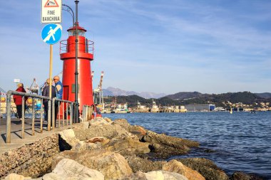 Bir körfezin sonundaki kırmızı fenerde insanlar geziniyor ve gün batımında arka planda bir liman var.