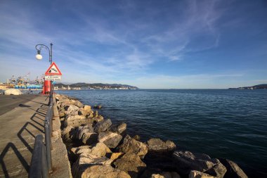 Bir körfezin sonundaki kırmızı fenerde insanlar geziniyor ve gün batımında arka planda bir liman var.