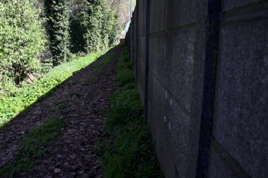 Yol, güneşli bir günde, bir ormandaki endüstriyel kompleksin beton bir sınır duvarıyla sınırlıydı.