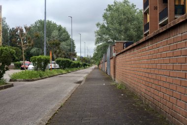 Bulutlu bir günde, bir kasabanın yerleşim bölgesinde ağaçlar ve çimenler olan cadde kaldırımdan görülüyor.